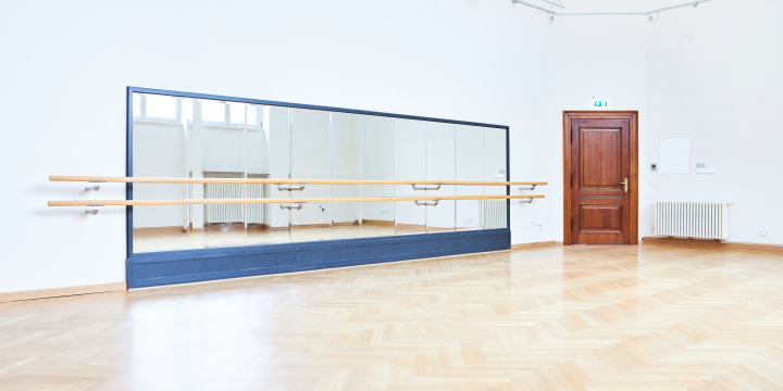 Eine Wand mit Spiegeln und Ballettstange, rechts davon eine braune Tür, ansonsten wirkt der Raum sehr hell und leer
