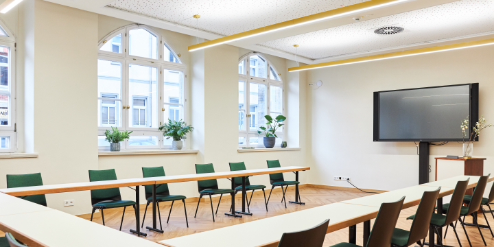 Heller Anna Auerbach Raum im Volkshaus Jena mit Konferenztisch in Hufeisenform und grünen Stühlen, mit Blick auf einen Monitor.