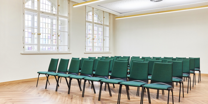 Vorderansicht auf fünf grün gepolsterter Stuhlreihen, links daneben zwei Buntglasfenster, in dem hellen Helene Holzman Raum des Volkshauses Jena.  ©JenaKultur, Karoline Krampitz